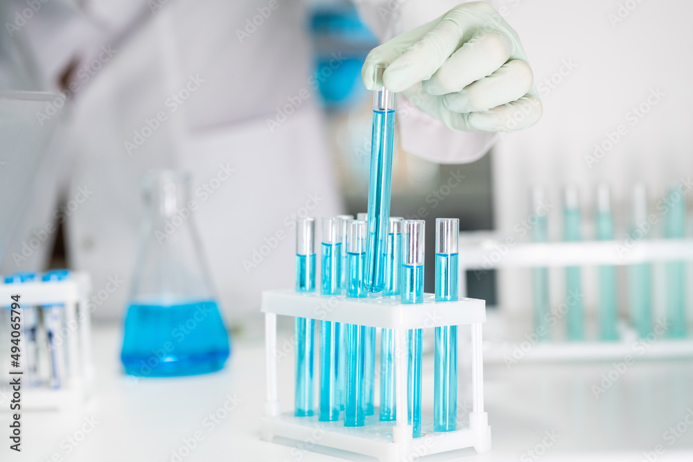 Gloved hand of female chemist or biotechnologist taking one of many flasks containing blue liquid chemical substance in laboratory Gloved hand of female chemist or biotechnologist taking one of many flasks containing blue liquid chemical substance in laboratory