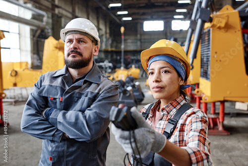 Two serious interracial workers of large factory looking at huge industrial machine while female technician regulating its height