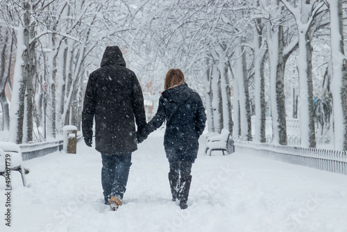 A couple with dark wear walking the snow with trees and chairs in both sides of the park