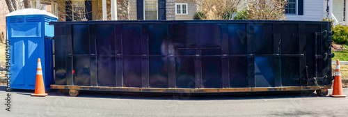 Black industrial dumpster container on neighborhood street with blue portable toilet. photo