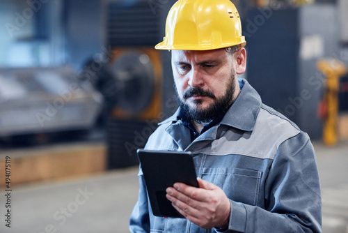 Mature bearded male worker of industrial plant in boilersuit and hardhat looking through data on screen of digital tablet © pressmaster