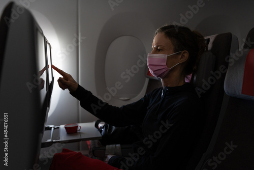 Woman with pink face mask interacting with touchscreen entertainment system sitting on airplane. Female passenger on plane during pandemic