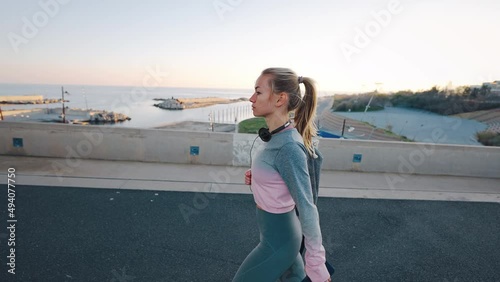 Focused confident young female athlete in sport suit walk along harbor at coastal city before start daily morning training outdoors hold cellphone and gym bag wear earphones around neck. Slow motion photo