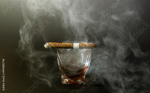 Whiskey drinks with cigars from Cuba Havana on black background. A glass of whiskey or cognac with a Smoking cigar lying on top. Front view.