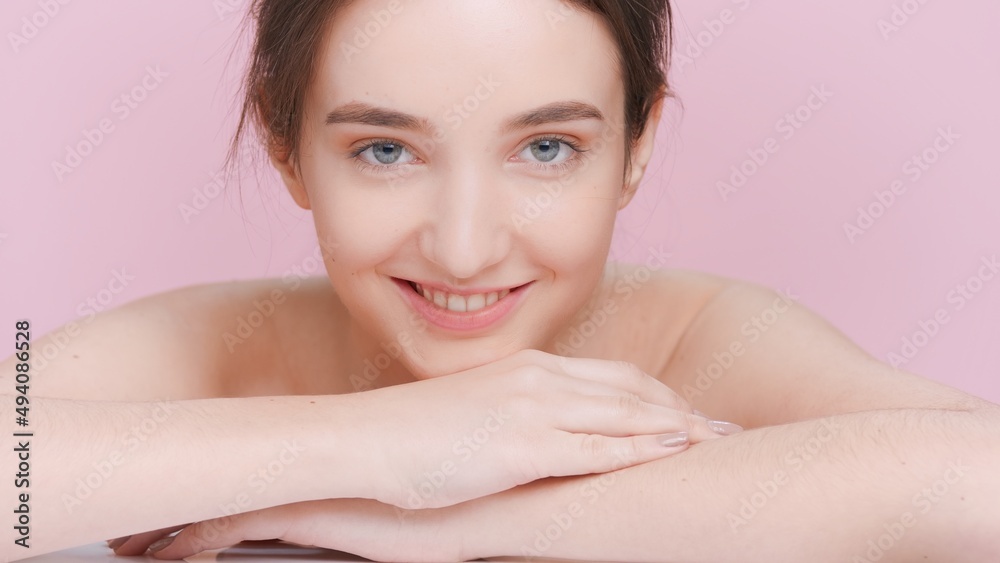 Young brunette woman slowly lays her chin on her hands, looks at the camera and smiles on pale pink background | Beauty and skincare product commercial concept. Young brunette woman slowly lays her chin on her hands, looks at the camera and smiles on pale pink background | Beauty and skincare product commercial concept.