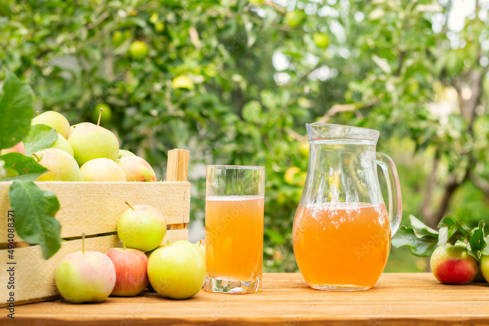 A box of apples and glass and pitcher of fresh apple juice on wooden table with garden background. Harvest of apples. Juice production A box of apples and glass and pitcher of fresh apple juice on wooden table with garden background. Harvest of apples. Juice production
