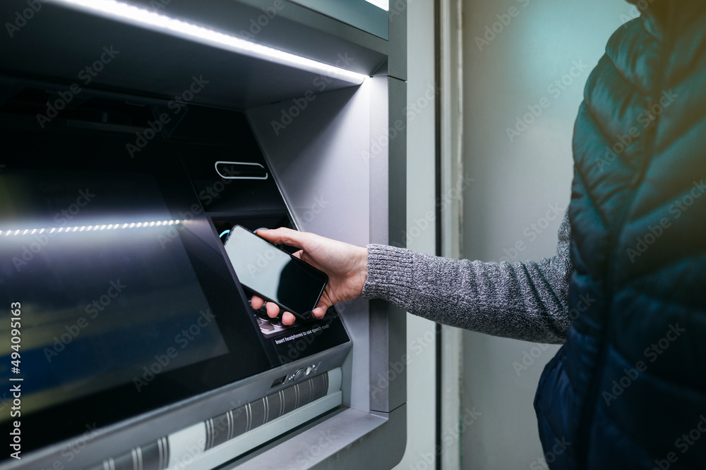 Man`s hand with an smartphone using an ATM street machine to withdraw some cash Man`s hand with an smartphone using an ATM street machine to withdraw some cash