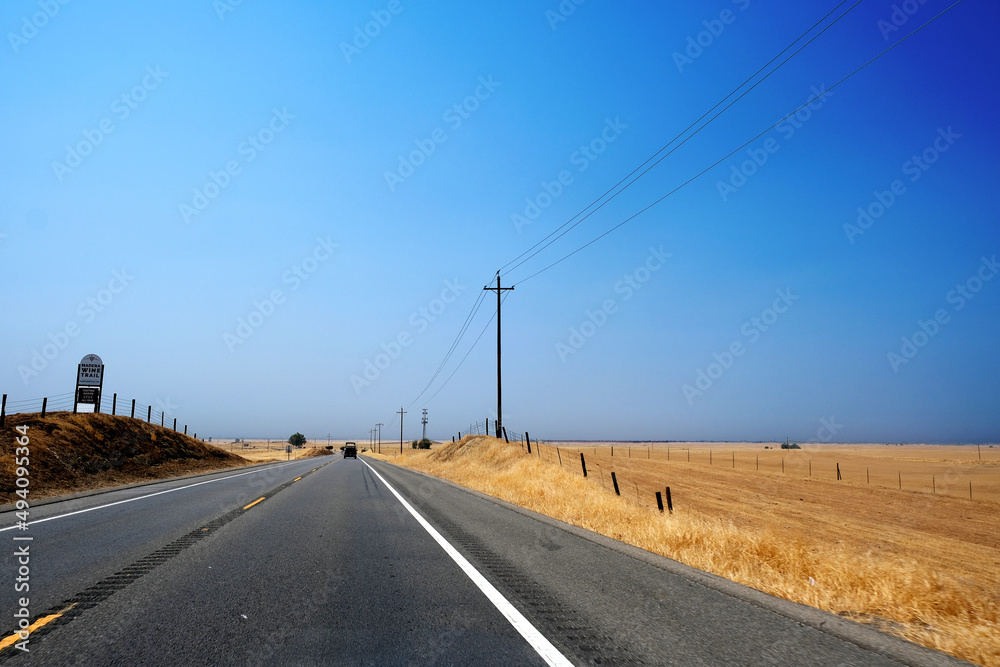 Lines separating mountains, hills, dried grass, farm land, and orchards beside highway 99 with wire fence under the blue sky