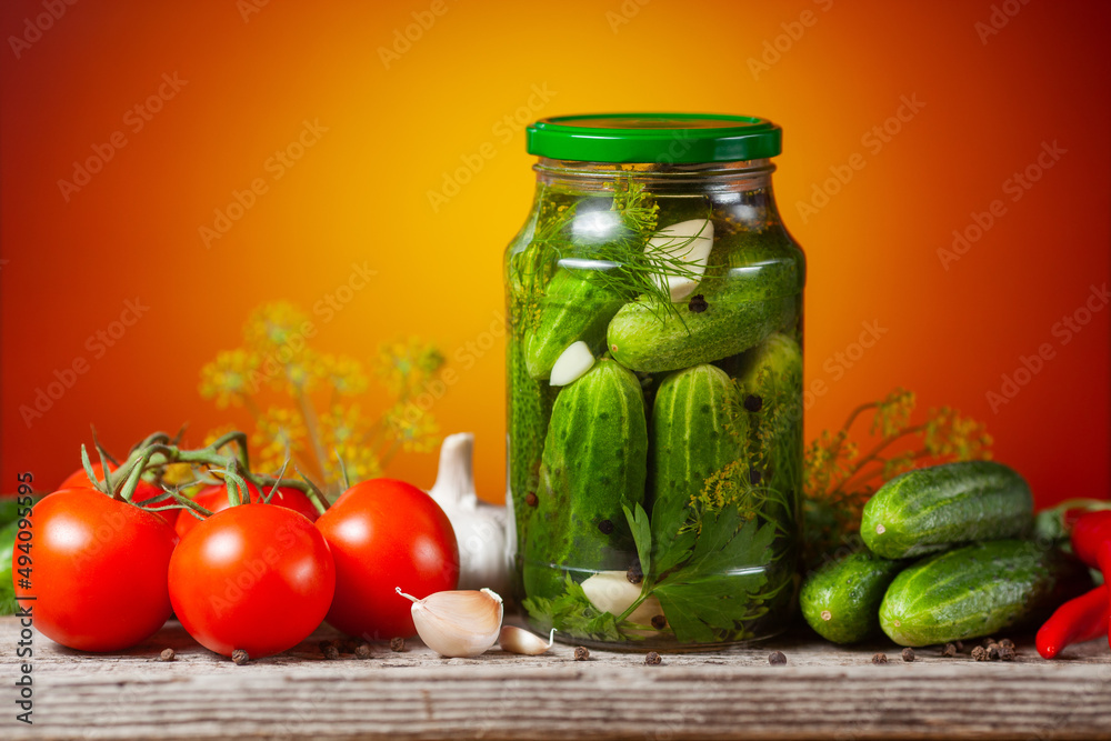 Pickled cucumbers in glass jars and spices and vegetables for preparation of pickles on orange background Pickled cucumbers in glass jars and spices and vegetables for preparation of pickles on orange background