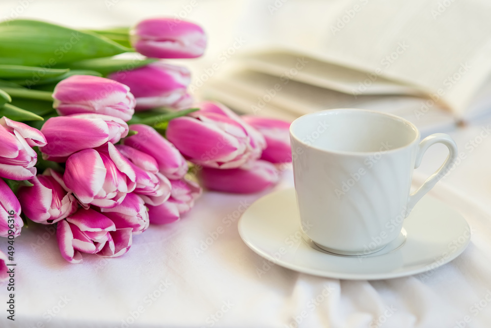 A bouquet of beautiful pink tulips lies on a white bed, next to a white cup and saucer and an open book A bouquet of beautiful pink tulips lies on a white bed, next to a white cup and saucer and an open book