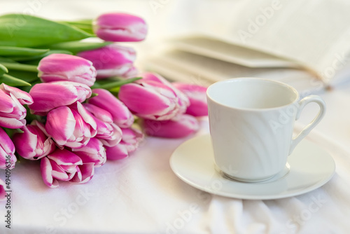A bouquet of beautiful pink tulips lies on a white bed, next to a white cup and saucer and an open book