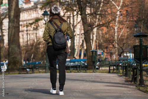 Close-up of a girl in a short jacket and sweatpants with a black bag on her back and a bottle of water walking along an asphalt road on a sunny day in early spring. Rear view horizontal orientation