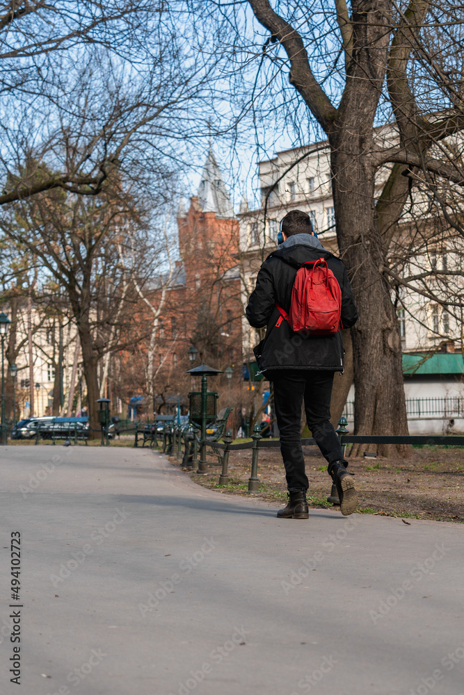 A young man with a red backpack on his back and headphones in a black jacket walks along the street of a big city on a sunny day in early spring. Back view
