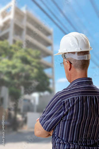 Man whith white hardhat on his head looking at building under construction.