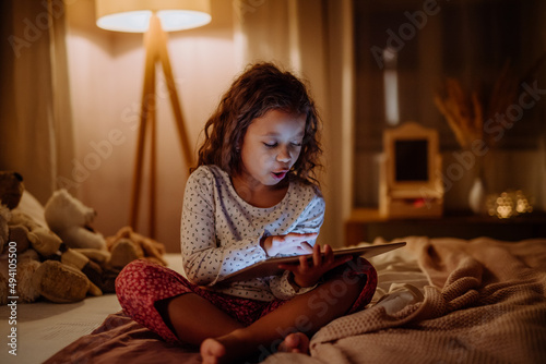 Happy little multiracial girl using tablet on bed in evening at home. photo