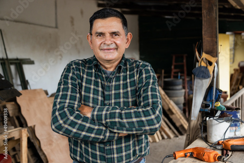 Hispanic carpenter smiling proud in wood shop of him - Proud owner of carpentry shop standing with arms crossed © Fernanda