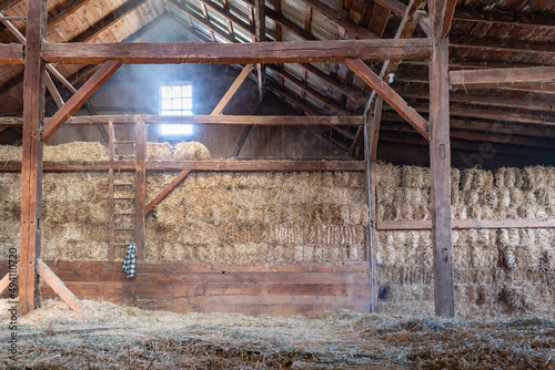Inside of Old Worn-Down Barn With Stacked Hay Bales Dark Wooden Beams and Light Shining Through Window