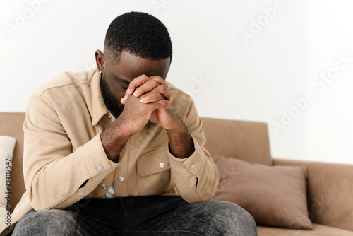 African American believing man sitting at home on sofa praying to God, asking for good luck, thoughtful bi-racial superstitious man holding hands in prayer, feeling gratitude, faith, religion concept