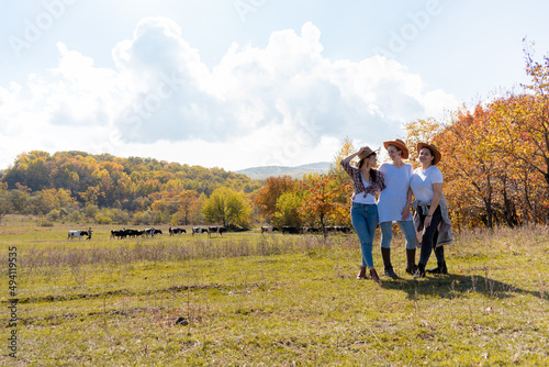 Three Diversity girlfriends in cowboy hats are standing in a meadow. Cows are grazing.