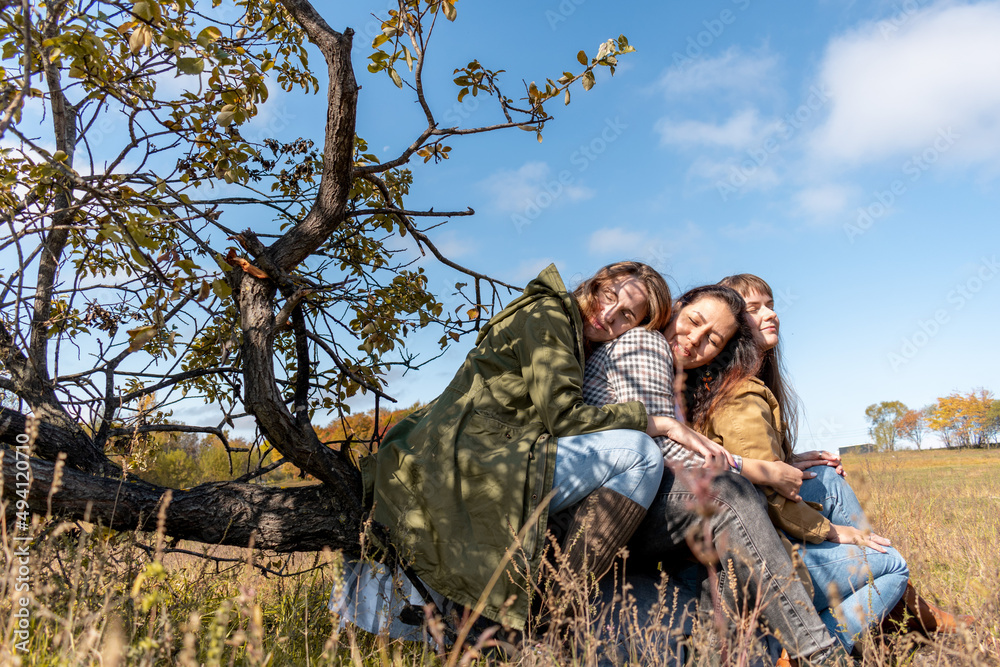 Three diverse female friends are hugging while sitting on a dry log in the autumn forest.