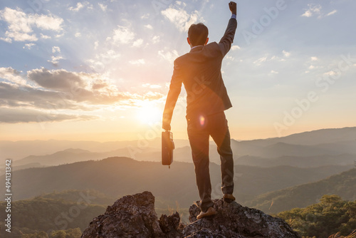 Businessman standing on a rock with his bag and raising his hand to show success