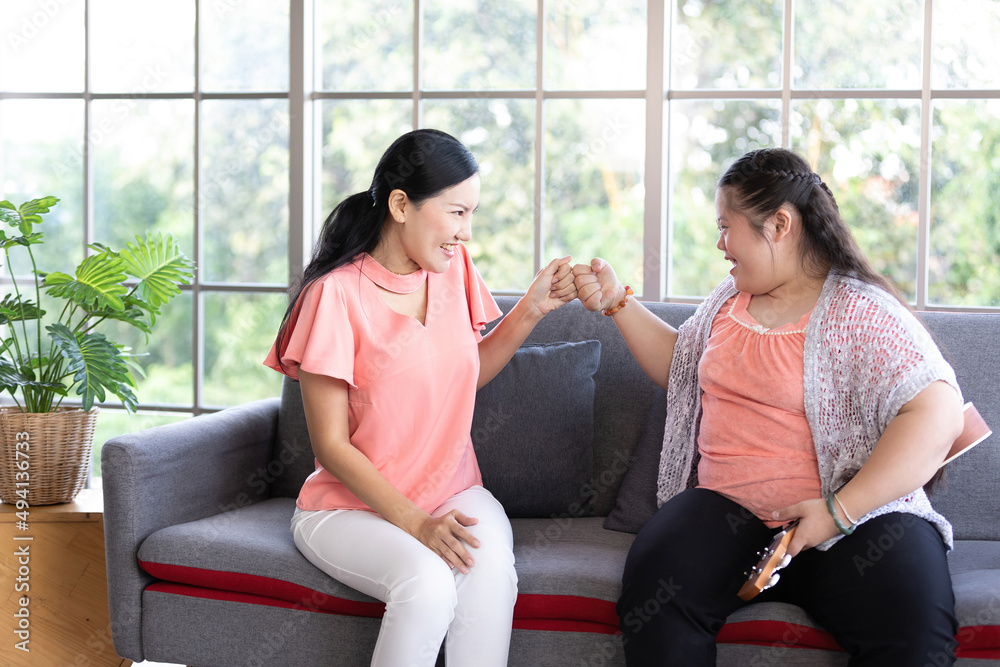 Obraz premium mother giving clashing fists to a girl with down syndrome, singing and playing ukulele or small guitar on sofa