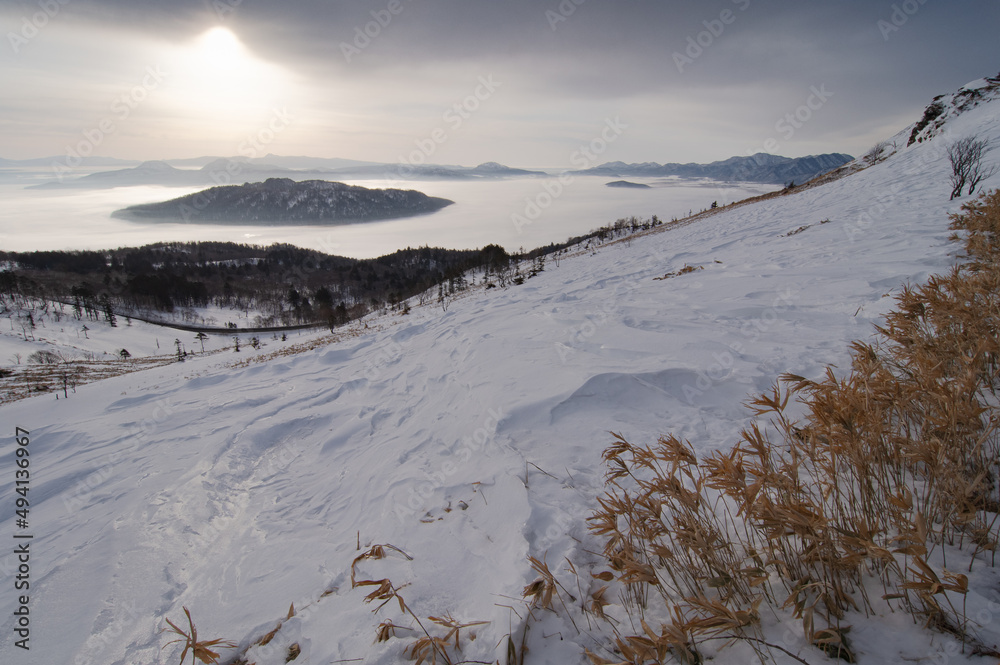 雪に覆われた斜面の向こうの靄に覆われた湖と曇り空に輝く太陽。日本の北海道の美幌峠の風景。