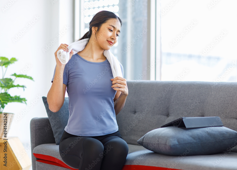 Obraz premium Asian young pretty happy healthy female athlete sport girl in casual sporty outfit sitting smiling on sofa holding using white towel cleaning drying sweat relaxing resting after exercising online