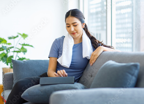 Asian young pretty happy healthy female athlete sport girl in casual sporty outfit sitting smiling on sofa holding using white towel cleaning drying sweat relaxing resting after exercising online