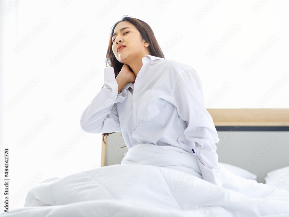 Asian young unhappy upset frowning face female teenager model in casual pajama outfit waking up in morning sitting under white blanket on bed in bedroom holding hands on neck having painful ache Asian young unhappy upset frowning face female teenager model in casual pajama outfit waking up in morning sitting under white blanket on bed in bedroom holding hands on neck having painful ache