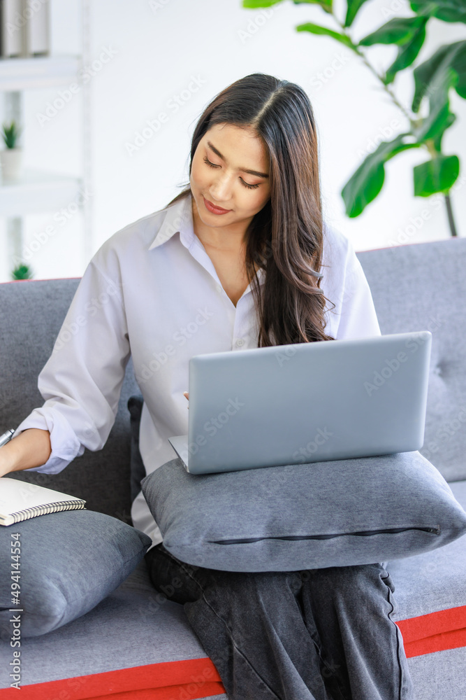 Asian young female professional successful businesswoman in casual outfit sitting on cozy sofa using pen writing note in notebook while working with laptop computer at home office in living room Asian young female professional successful businesswoman in casual outfit sitting on cozy sofa using pen writing note in notebook while working with laptop computer at home office in living room