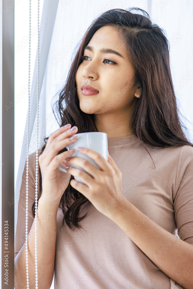 Asian young beautiful female housewife in casual outfit standing smiling looking outside view through glass window in living room holding drinking hot coffee from white mug after waking up in morning Asian young beautiful female housewife in casual outfit standing smiling looking outside view through glass window in living room holding drinking hot coffee from white mug after waking up in morning