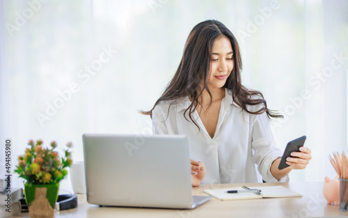 Asian young beautiful happy professional successful businesswoman designer sitting smiling at workstation desk using laptop notebook computer and smartphone working remotely online at home office