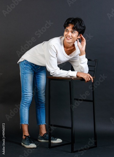 Portrait studio full body shot of Asian young LGBTQ gay glamour handsome male model in casual white shirt jeans sneakers sitting on tall chair laughing holding hand cover mouth on black background