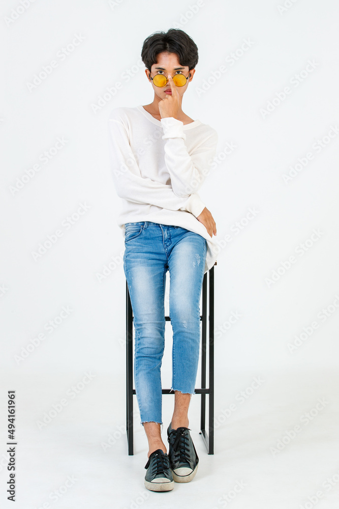 Portrait studio shot of Asian young LGBT gay happy bisexual homosexual male fashionable model in casual outfit fashion sunglasses standing leaning at tall chair smiling posing on white background