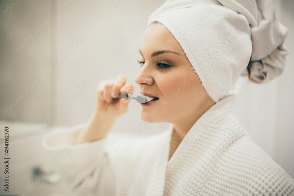 Young woman wearing white bathrobe and tower on head brushing her teeth using toothbrush and paste. Young woman wearing white bathrobe and tower on head brushing her teeth using toothbrush and paste.