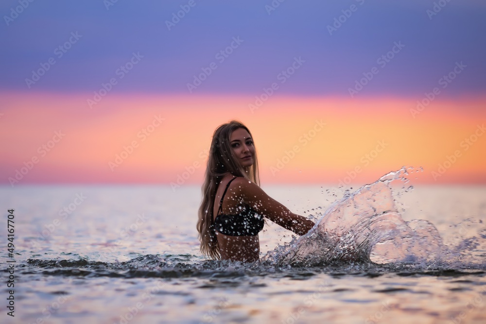 A girl with blond hair in a black swimsuit splashes to the sides while sitting in an estuary on a sunset background