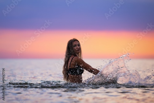 A girl with blond hair in a black swimsuit splashes to the sides while sitting in an estuary on a sunset background