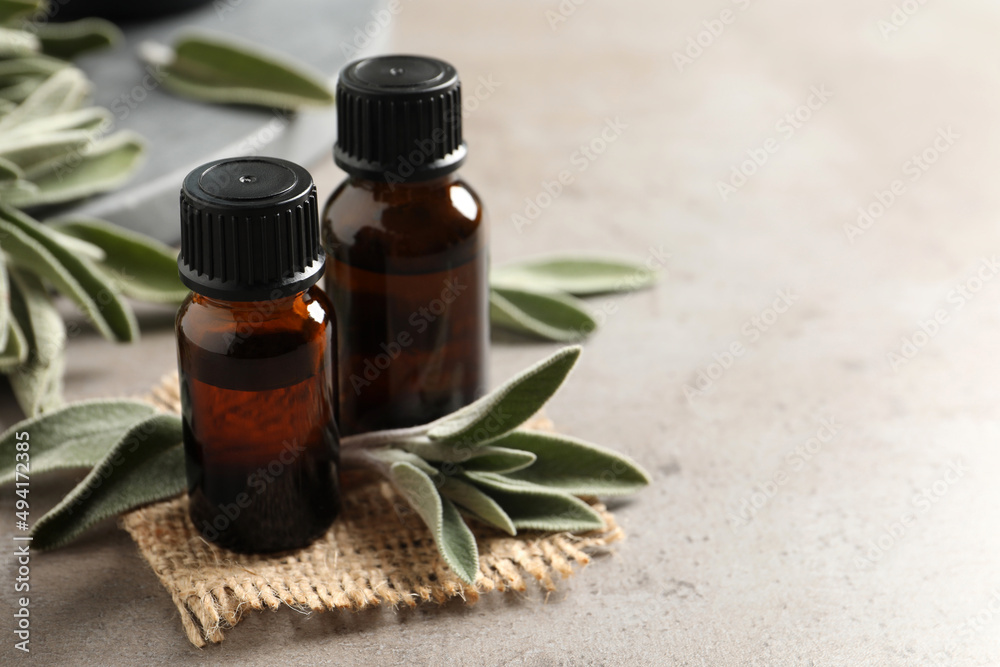 Bottles of essential sage oil, twigs and leaves on light grey table. Space for text