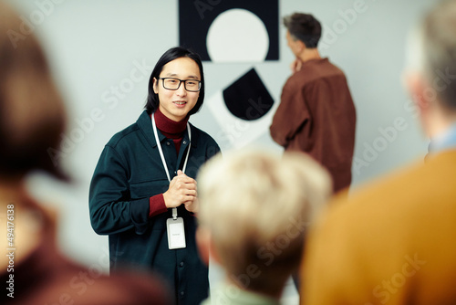 Young Asian man working in modern arts gallery standing in front of group of unrecognizable people speaking about exhibition