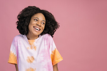 Beautiful smiling African woman looking away isolated on pink background, copy space. Happy curly haired fashion model wearing stylish tie dye t shirt posing for pictures in studio 
