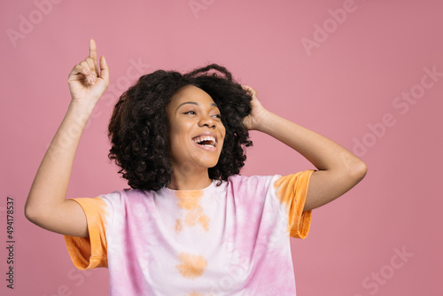 Smiling African American woman dancing looking away isolated on pink background. Happy curly haired teenager wearing stylish tie dye t shirt having fun 