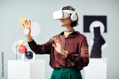 Young Caucasian woman wearing VR headset looking at art objects while visiting modern exhibition using augmented reality technology photo