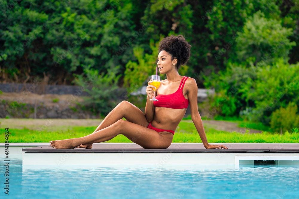 A young African American woman in a red bikini sunbathes by the pool while sipping a non-alcoholic fruit cocktail
