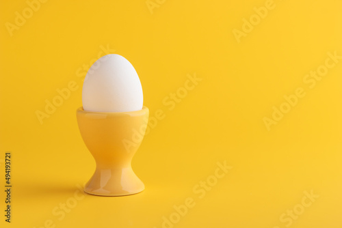 White fresh egg in the yellow egg stand, cup for breakfast on solid yellow background, vertical with empty space, Easter holiday, healthy eating