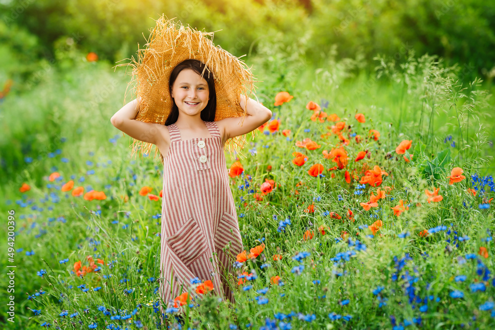 Happy little girl in a summer dress and straw hat among wildflowers. Lots of poppies and cornflowers in the green grass. Happy little girl in a summer dress and straw hat among wildflowers. Lots of poppies and cornflowers in the green grass.