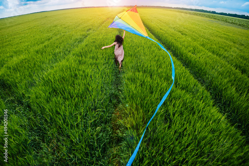 Little happy girl running with kate in hands on green wheat field. Large colored rainbow kite with long blue tail. Top back view. photo