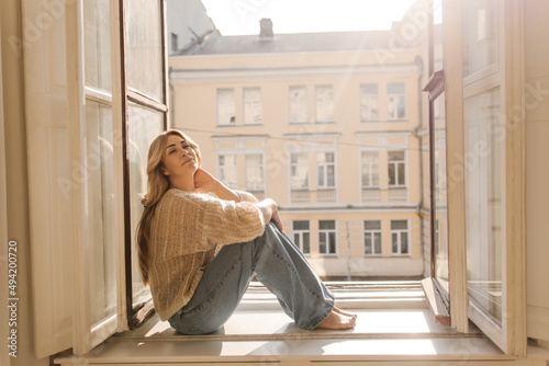 Beautiful adult caucasian woman is sitting on windowsill of house near open window overlooking sunny city. Blonde woman in sweater and jeans looks at camera. Vacation concept