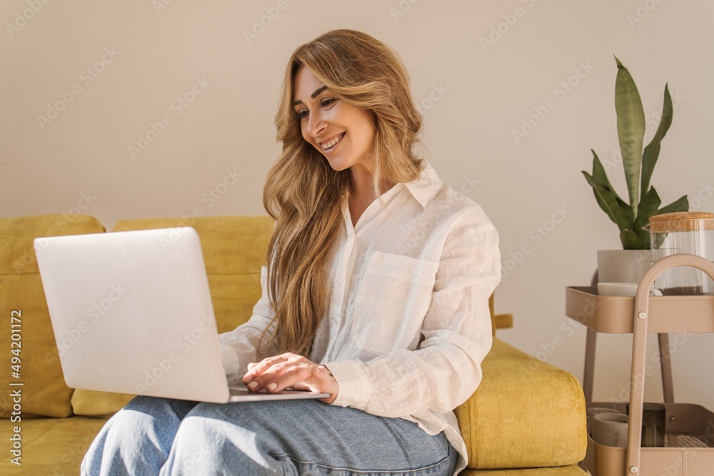 Adult fair-skinned business blonde uses notebook at home while sitting on comfortable sofa. Smiling lady is wearing shirt and jeans. Remote work concept Adult fair-skinned business blonde uses notebook at home while sitting on comfortable sofa. Smiling lady is wearing shirt and jeans. Remote work concept