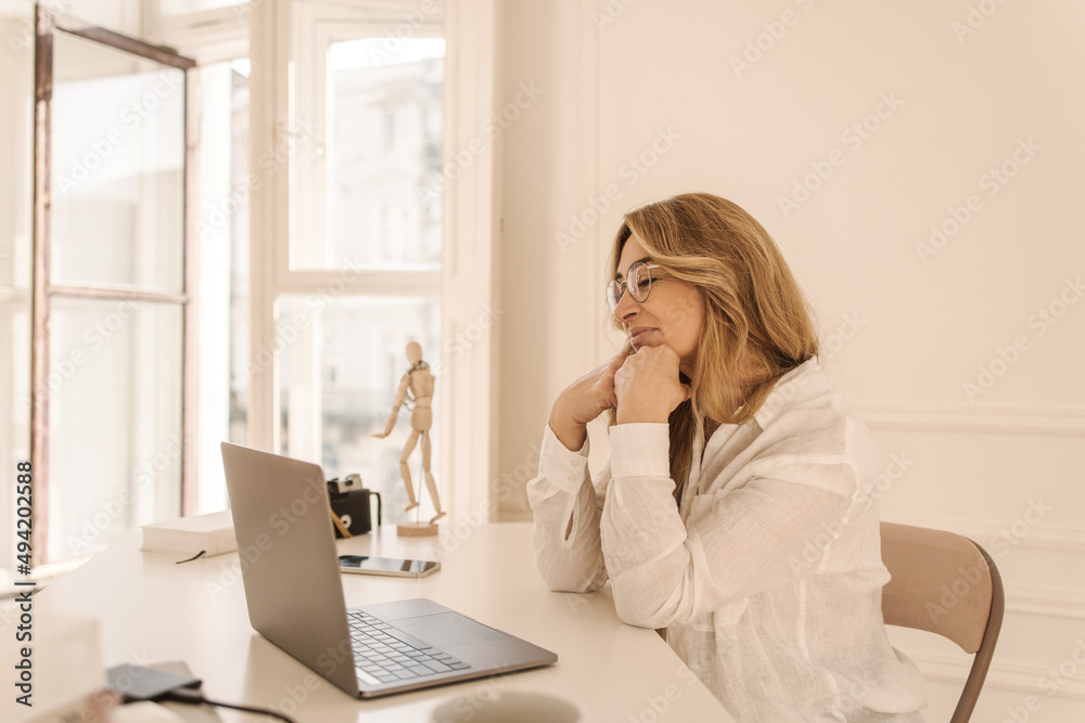 Talented adult caucasian woman sits in front of notebook and writes her new book in lightroom. Writer with blond hair is looking for inspiration. Work from home concept Talented adult caucasian woman sits in front of notebook and writes her new book in lightroom. Writer with blond hair is looking for inspiration. Work from home concept